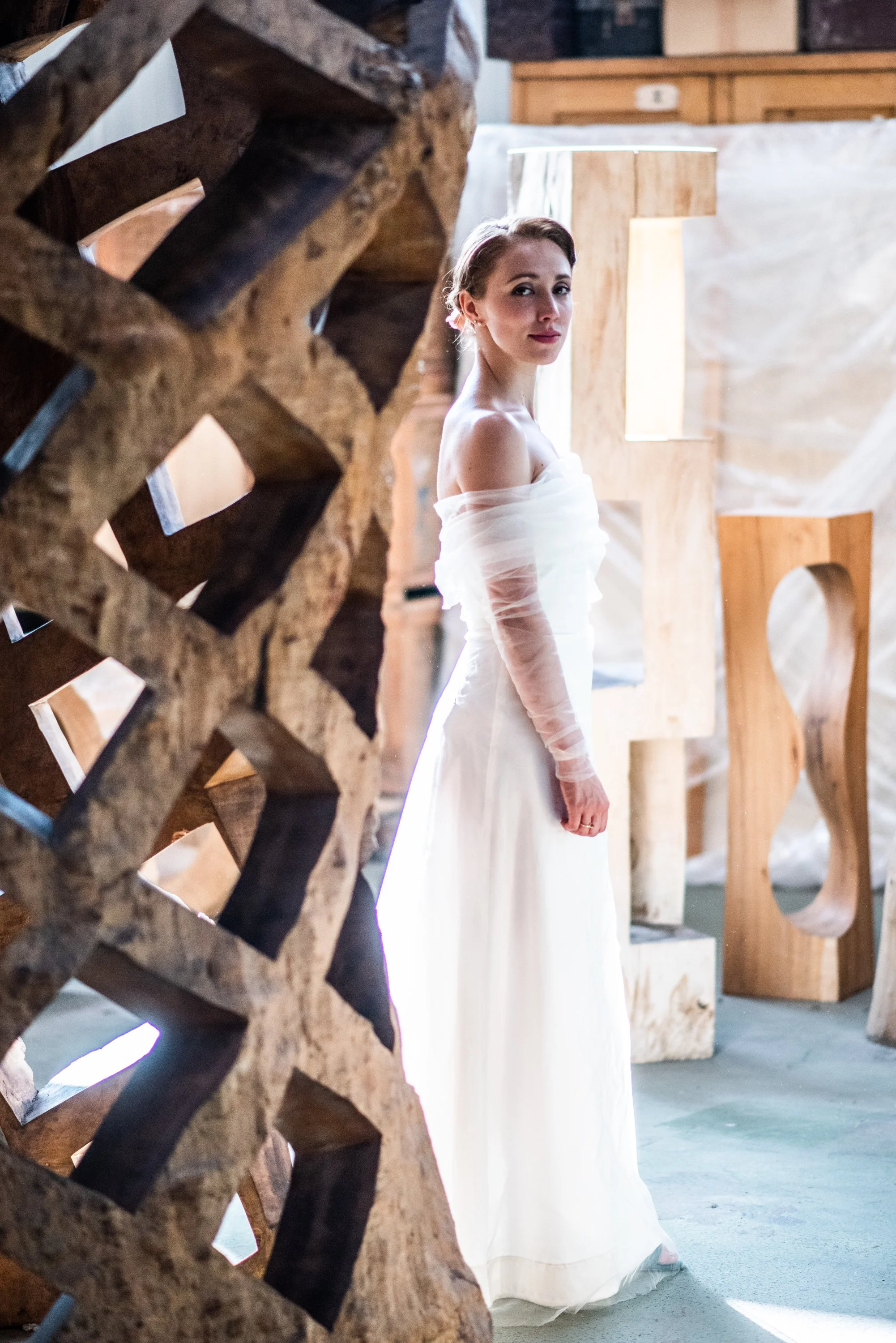 A woman in a white strapless dress stands in a rustic setting with wooden sculptures and logs, looking at the camera.