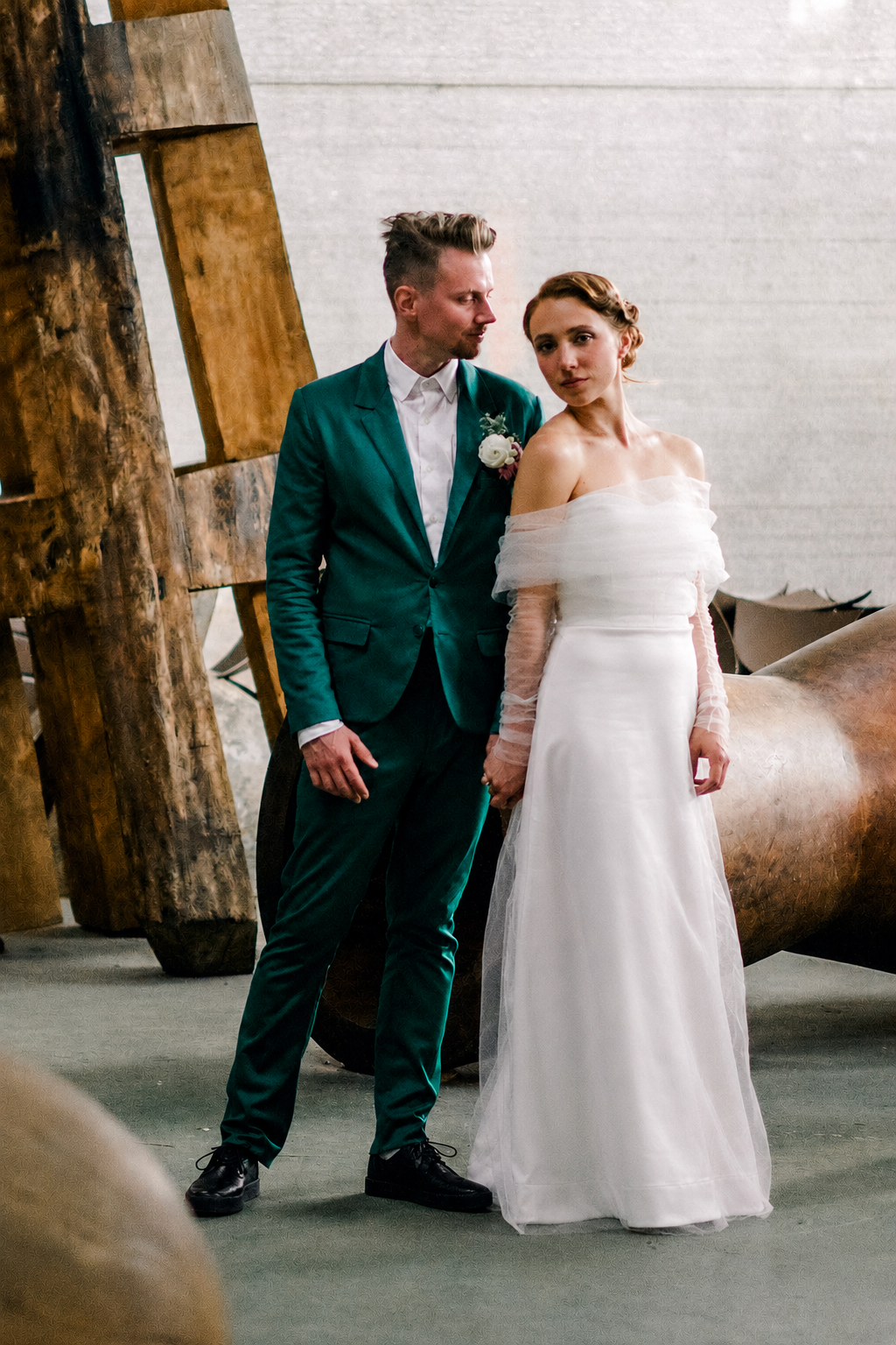 A bride and groom holding hands during their wedding ceremony, standing indoors near rustic wooden decor, with the groom in a teal suit and the bride in an off-shoulder white wedding dress.