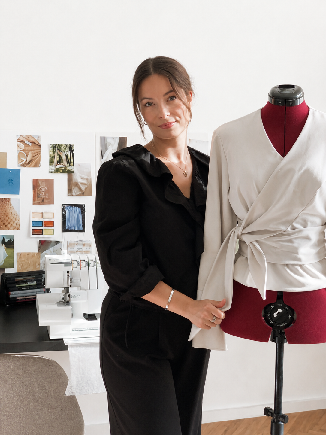 Woman standing beside a dress mannequin with a white wrap top and tied waist, in a fashion designer's workspace.