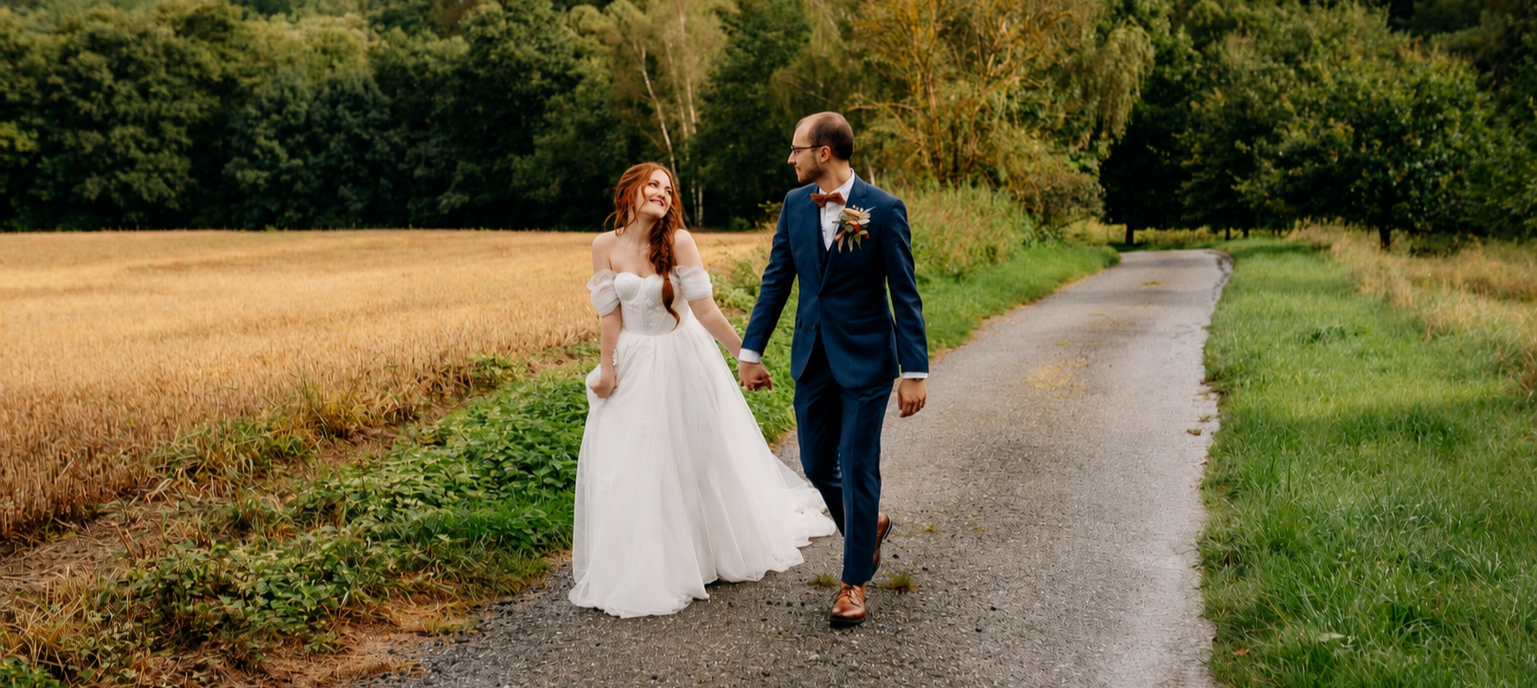 A couple in wedding attire walking hand in hand on a gravel path through a rural landscape, surrounded by green trees and fields.