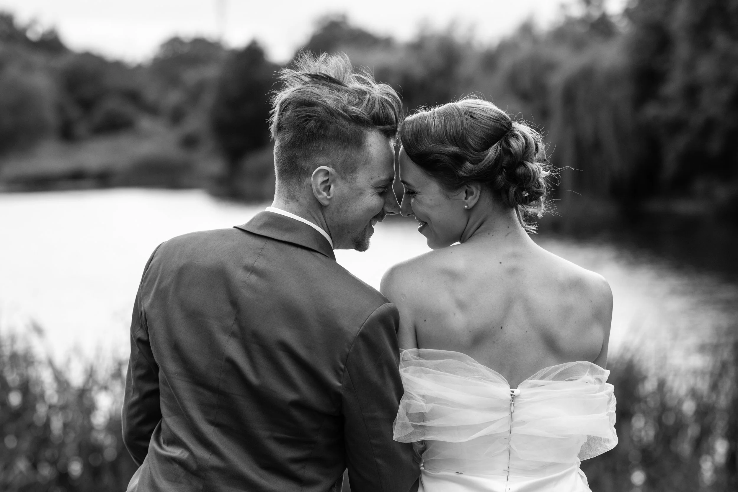 A black and white photograph of a couple smiling with their foreheads touching, outdoors near a body of water with blurred trees in the background.