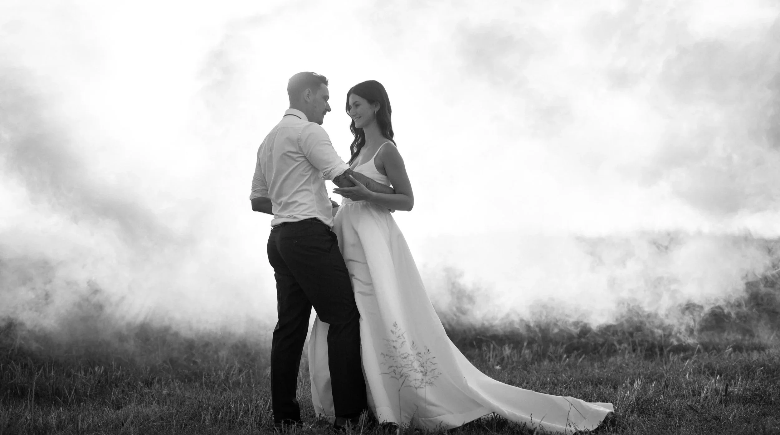 A black and white photo of a couple on their wedding day, standing outdoors on a grassy field, holding hands and smiling at each other with overcast clouds in the background.