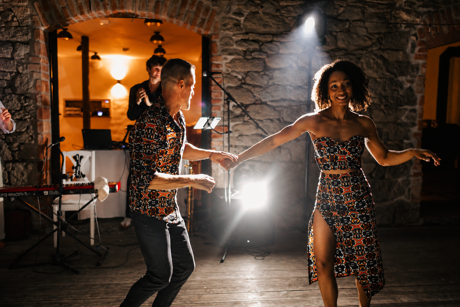 A couple dancing hand in hand in a warmly lit venue with stone walls and a wooden floor. The woman is smiling, wearing a strapless dress with a high slit. The man is dressed in a colorful, patterned shirt and dark pants. A musician with a laptop and microphone is in the background, with another person clapping nearby.