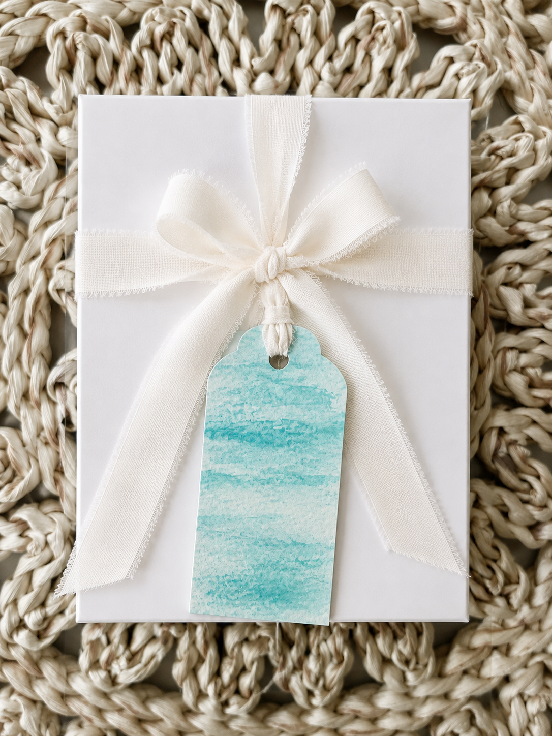 A white gift box wrapped with a cream-colored ribbon tied in a bow, featuring a blue watercolor gift tag attached, placed on a textured woven mat.
