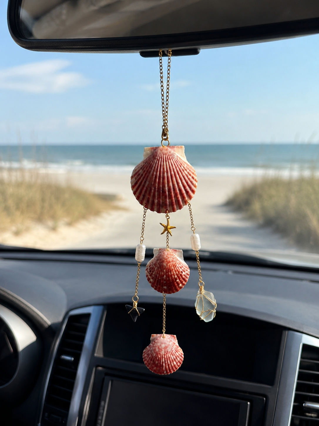 A seashell and starfish hanging decoration inside a car, with a view of a beach and ocean through the windshield.