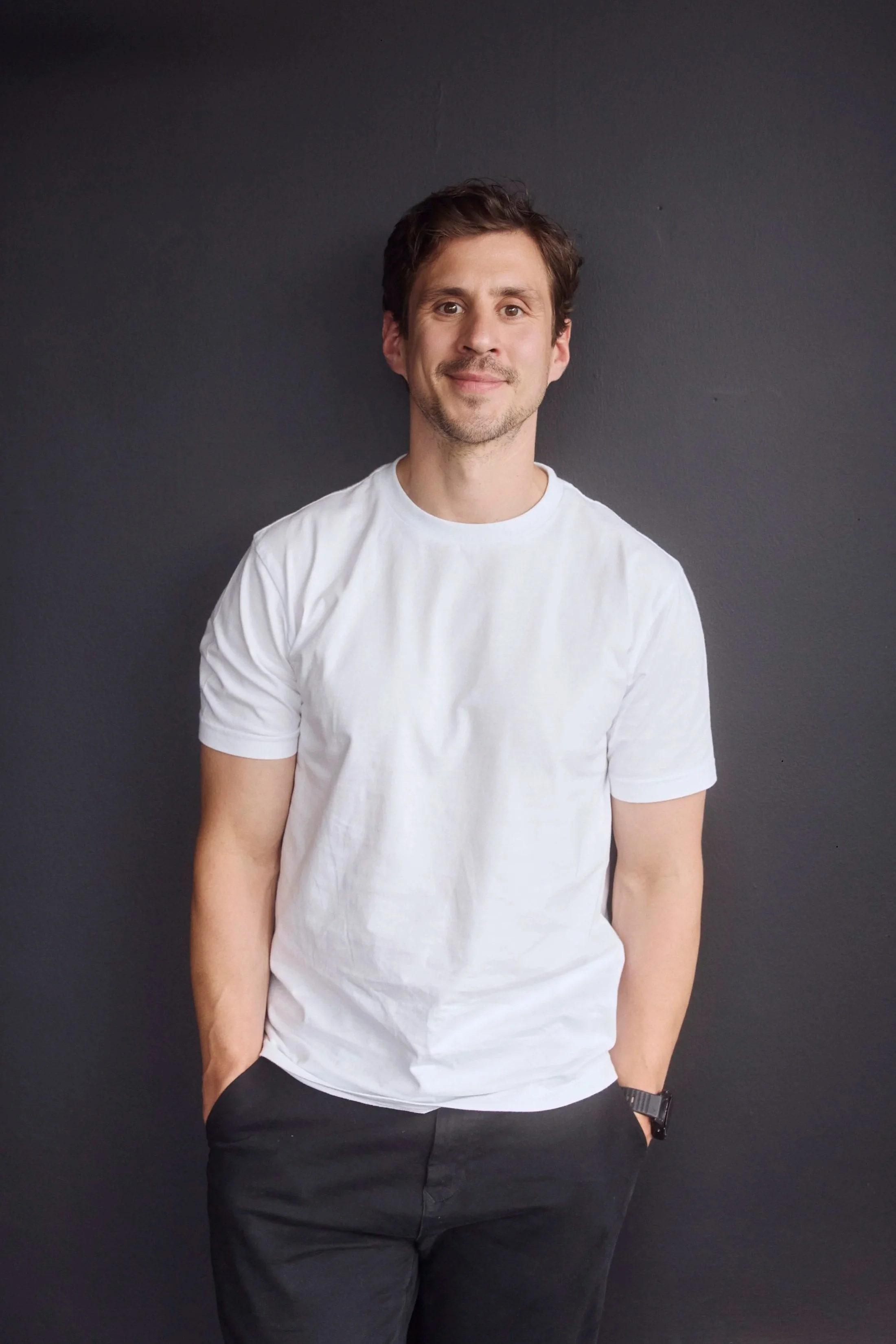 A young man with short brown hair and a slight beard, wearing a white t-shirt, black pants, and a watch on his left wrist, stands against a plain dark background. He has a relaxed posture with one hand in his pocket and a slight smile.