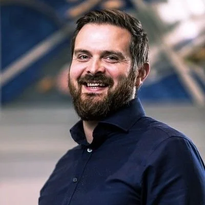 A smiling man with a beard and dark hair, wearing a dark blue shirt, standing outdoors with a blurred background.