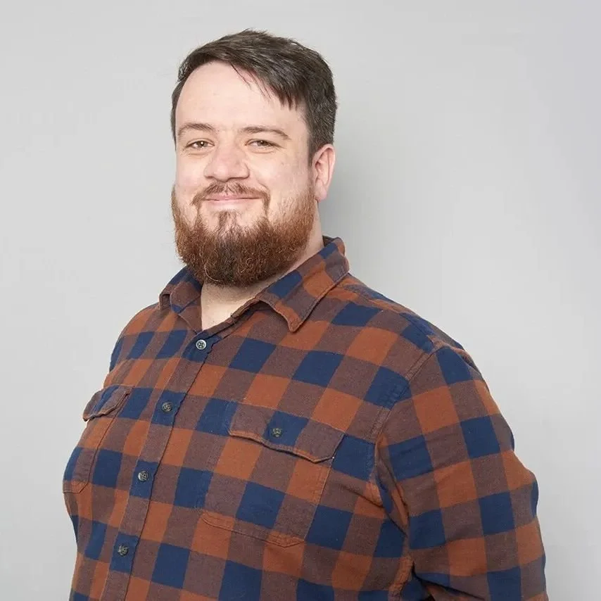 A man with short dark hair and a beard, smiling, wearing a brown and blue checkered shirt, standing against a plain light gray background.