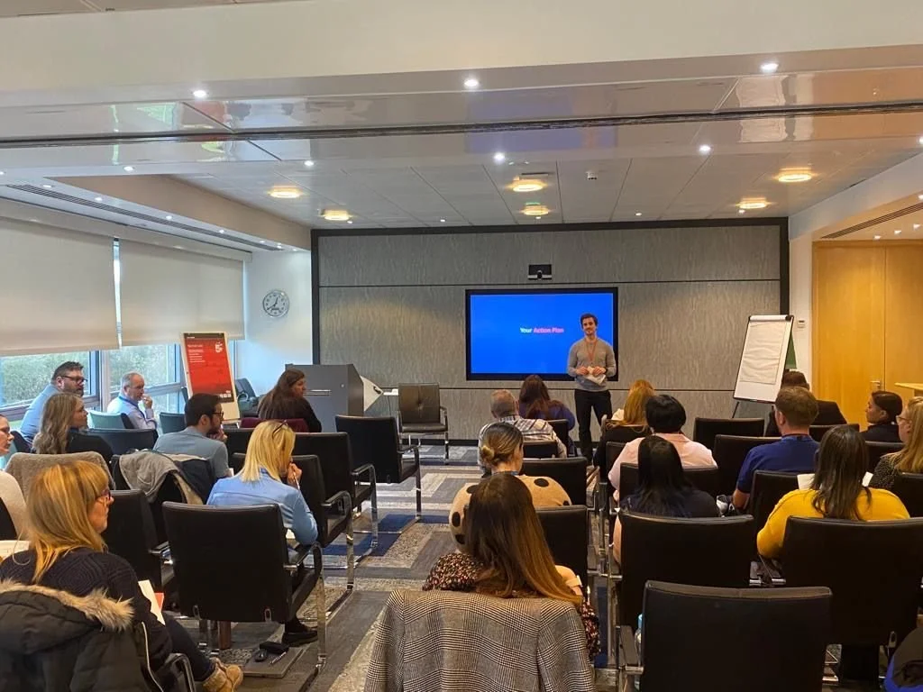 A group of people attending a presentation in a conference room with a presenter standing in front of a large screen.