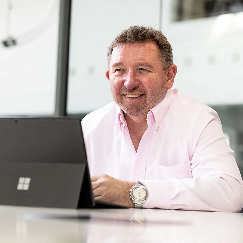 A man with short brown hair, a beard, and a pink dress shirt smiling while sitting at a table with a closed Microsoft Surface tablet in front of him. He is wearing a silver watch on his left wrist.