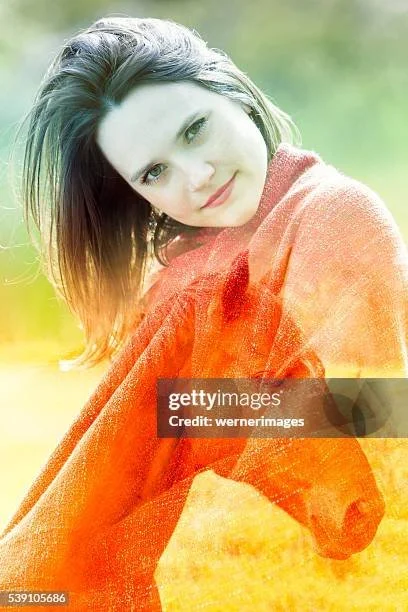 A woman with shoulder-length brown hair smiling and wrapped in an orange blanket outdoors, with a blurred green background.