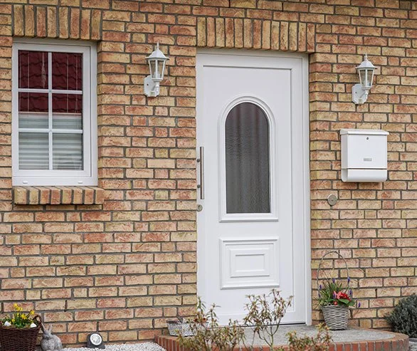 Exterior brick house with a white front door, a window with shutters, two wall-mounted lantern lights, a mailbox, and potted plants.
