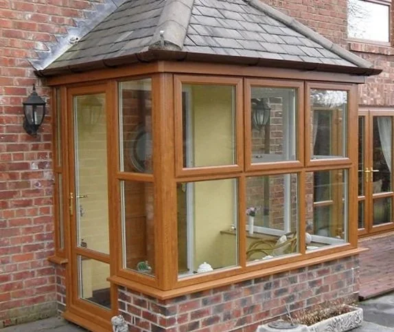 A wooden-framed glass conservatory extension attached to a brick house, with a shingled roof and a door leading inside.