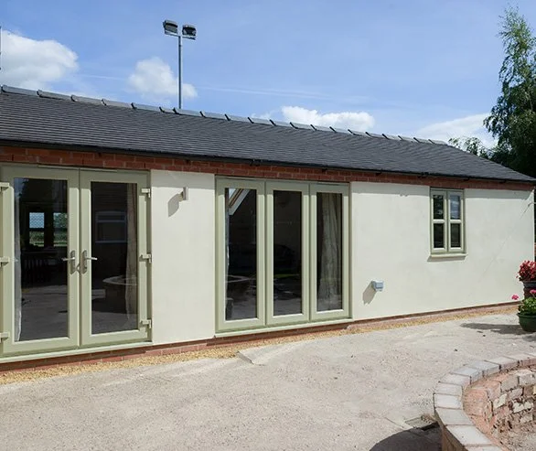 A single-story house with a gray roof and beige exterior walls, featuring glass doors and small windows, with a small courtyard and potted plants outside.