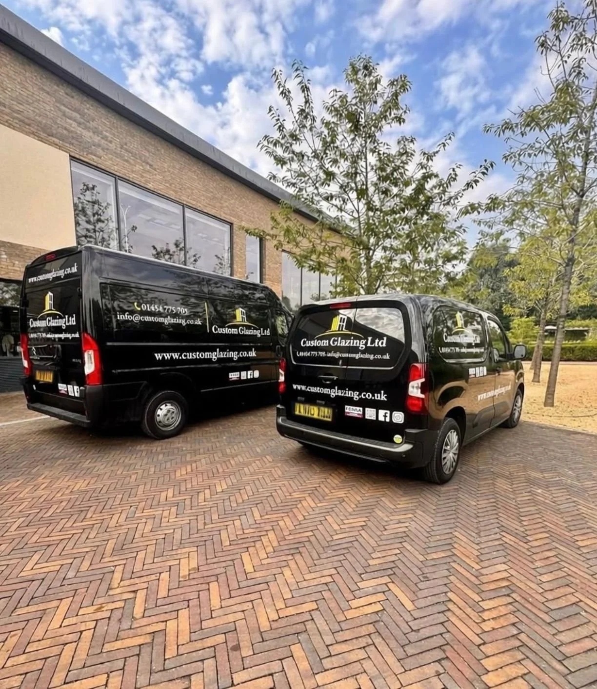 Two black vans with branding from Custom Glazing Ltd parked outside a modern brick building with large windows, surrounded by trees and a brick-paved parking lot on a partly cloudy day.