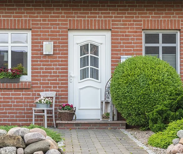 Brick house entrance with white door, two windows with flower boxes, a small outdoor chair with flowers, and well-maintained bushes and rocks in the front yard.