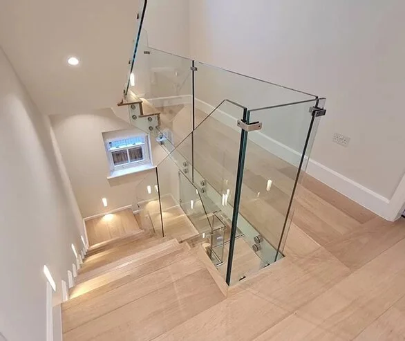 View of a modern staircase with wooden steps, glass railings, and a small window at the landing, taken from the top looking down.