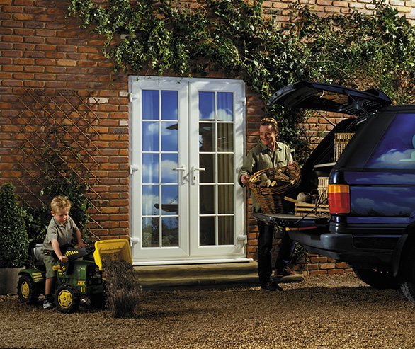 A man and a child outdoors near a brick house with double glass doors, a garden, and a car with its trunk open. The child is sitting on a small green toy tractor, and the man is placing an animal, possibly a large cat, into the trunk of the car.