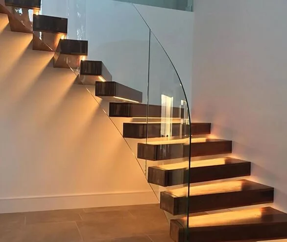 Modern floating staircase with wooden steps, glass railing, and warm lighting on the steps, next to a plain wall in a contemporary interior.