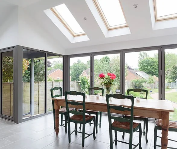 Sunlit dining room with large glass sliding doors, skylights, and a wooden table with six green chairs and a vase of pink flowers.