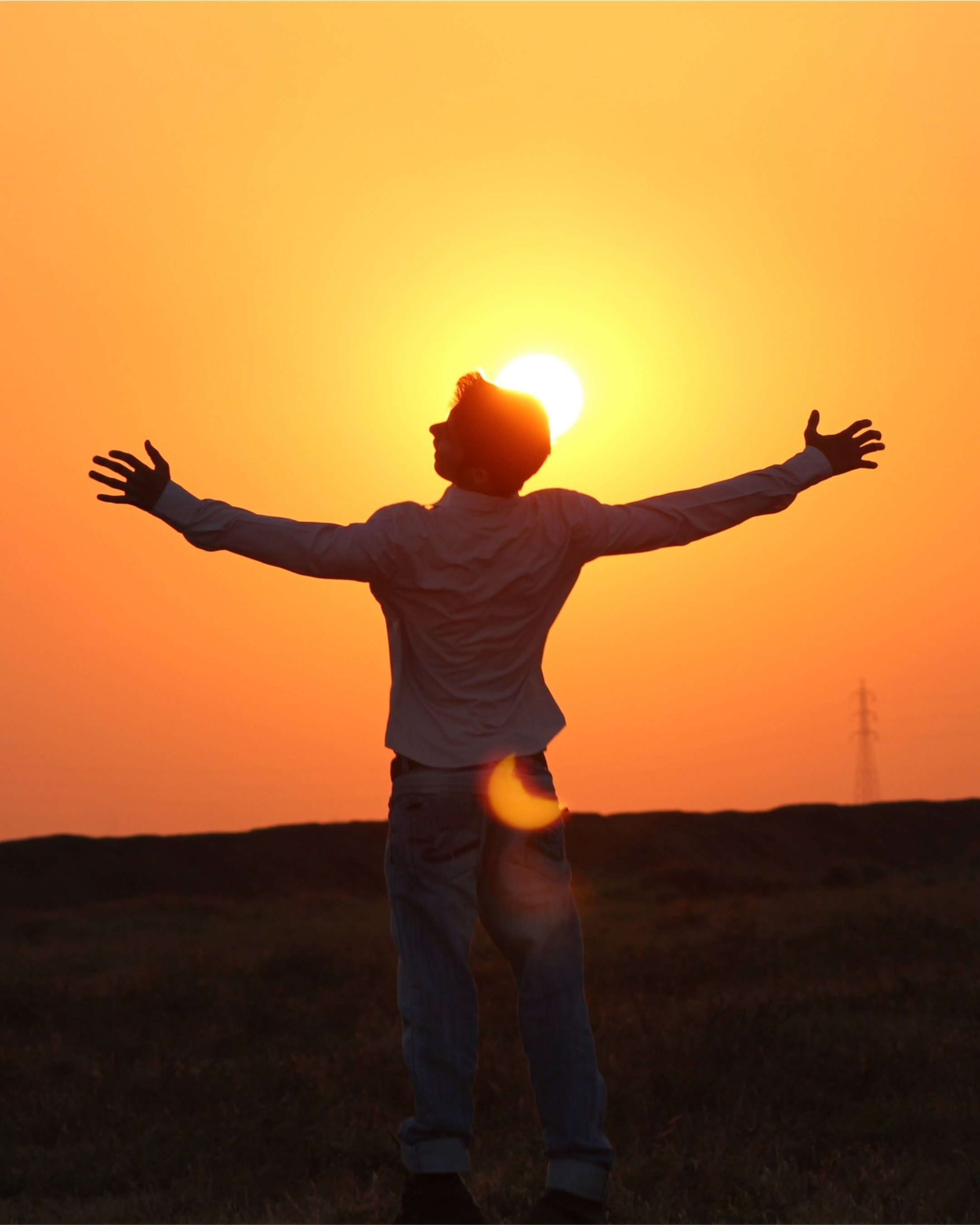 Person standing outdoors during sunset with arms outstretched, facing the sun, silhouetted against the orange sky.