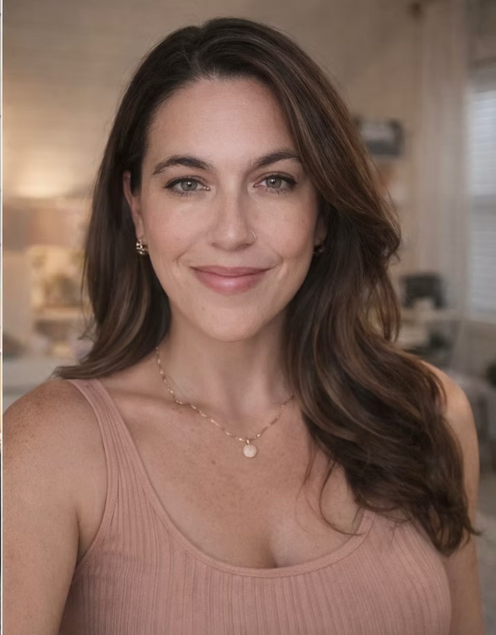A woman with long brown hair, wearing a pink tank top, smiling in a warmly lit room with blurred background.