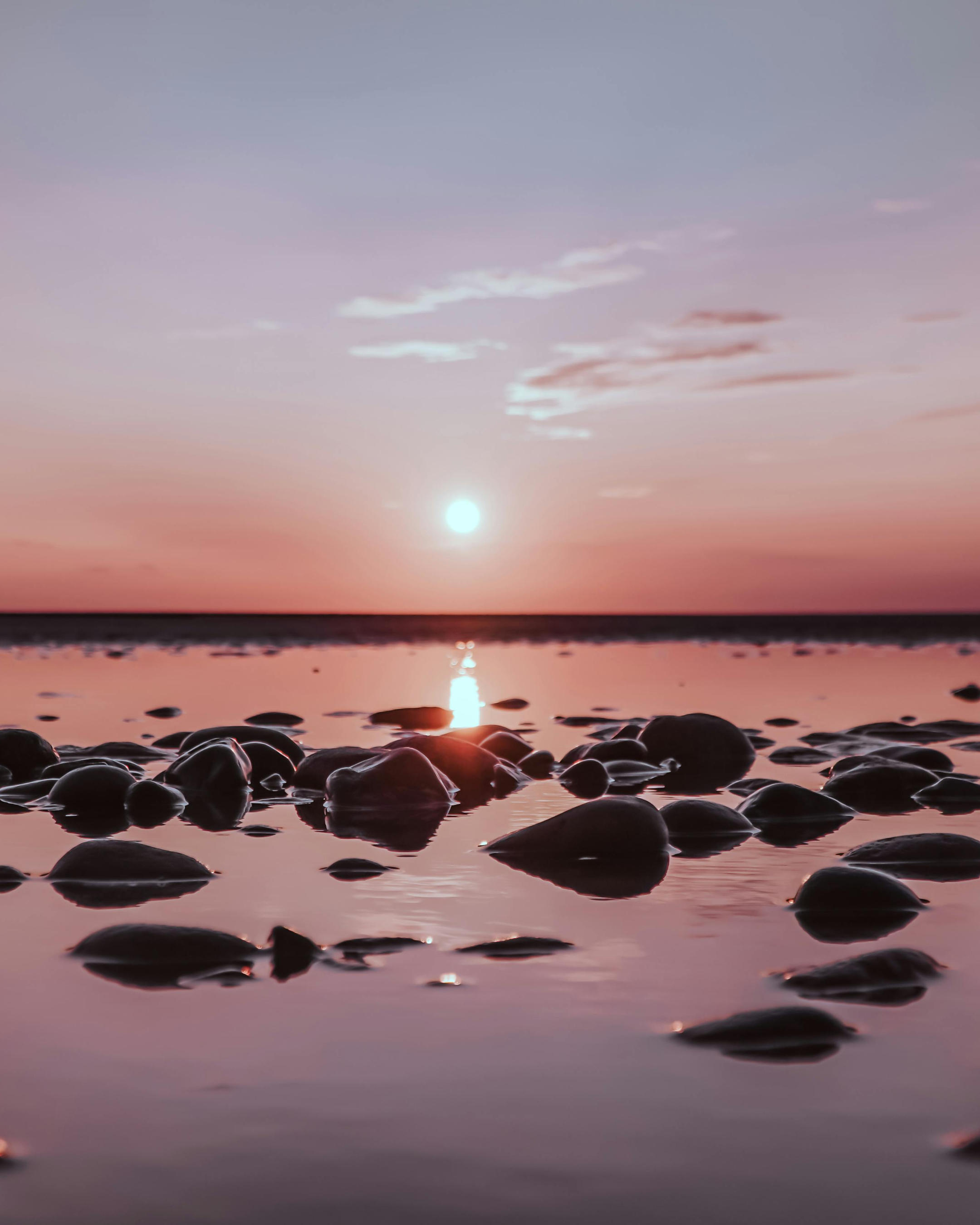 Sun setting over a calm body of water with rocks in the foreground and a pinkish-purple sky.