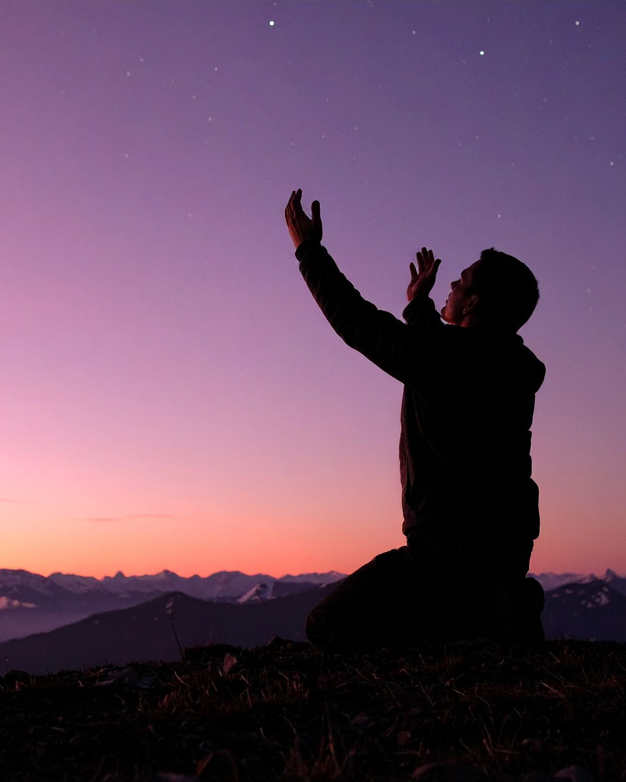 Silhouette of a person kneeling and reaching towards the sky during a sunset or twilight, with purple and pink hues in the sky and mountain ranges in the background.