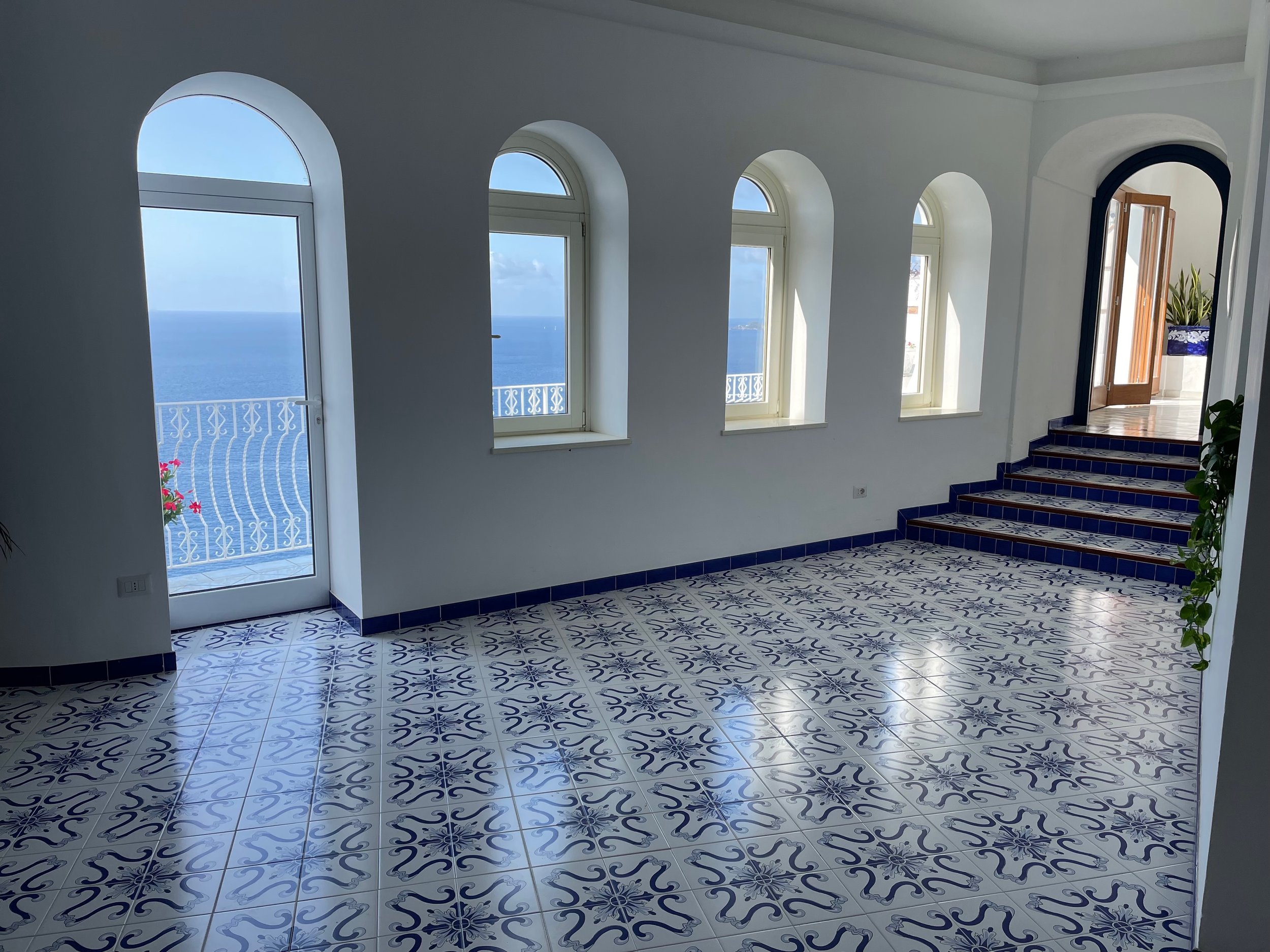 Interior view of a room with four arched windows showing a seascape, staircase with patterned blue tiles, and a door leading to a balcony with ocean view.