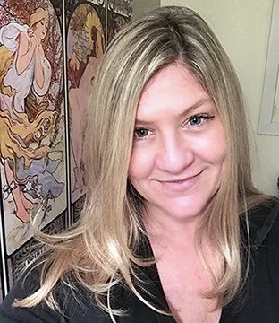 A blonde woman smiling, with Indian-inspired artwork in the background.