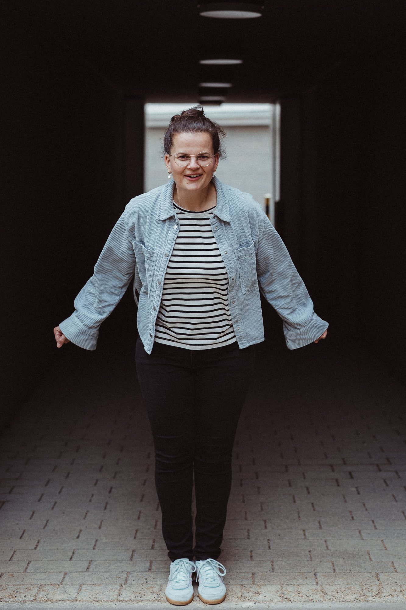 Eine lachende Frau mit Brille, gestreiftem Oberteil, Jeansjacke, schwarzen Hosen und weißen Turnschuhen, die nach vorne läuft in einem dunklen Tunnel.