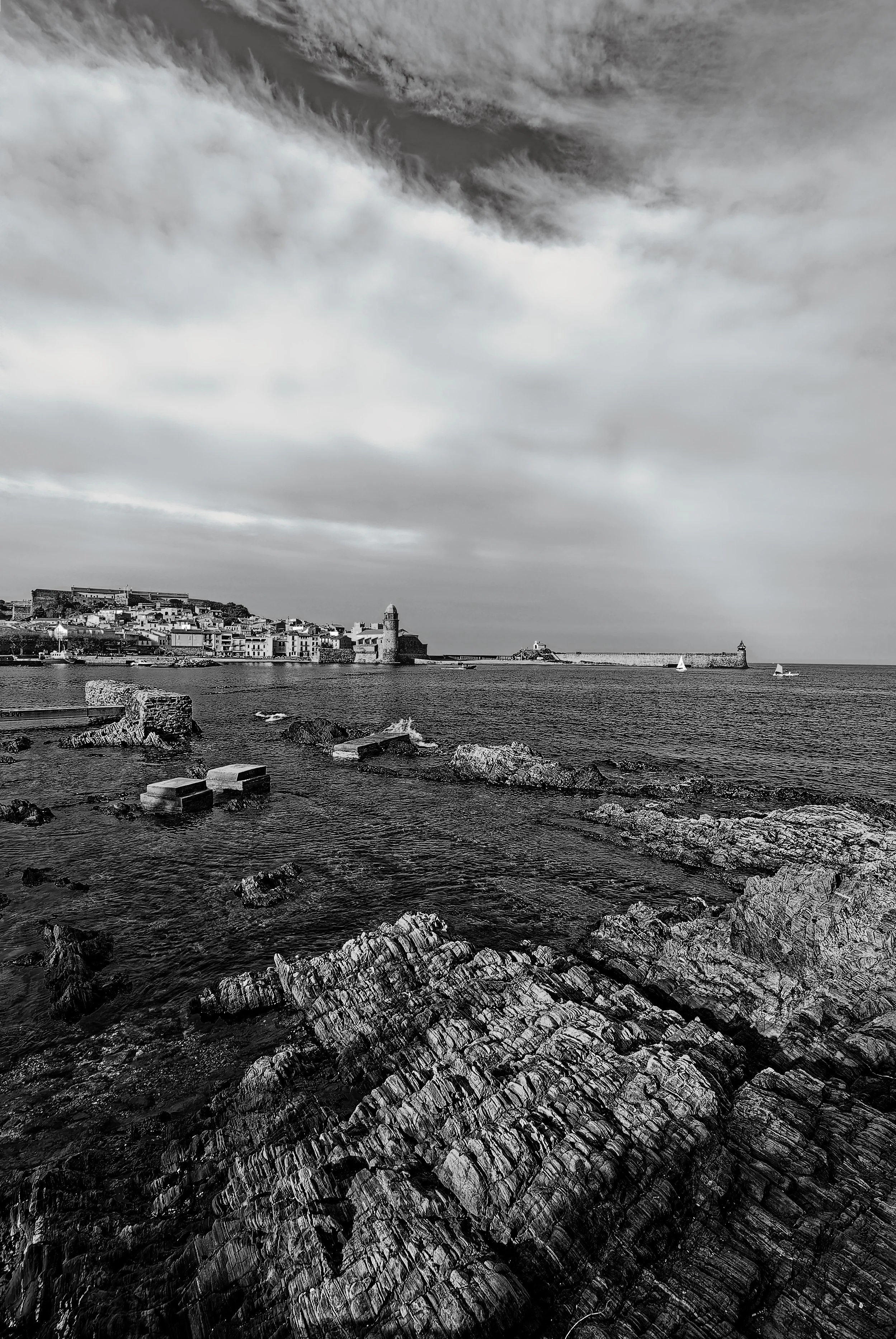 A black and white photo of a coastal scene featuring a rocky shoreline in the foreground, a body of water with a few sailboats, a historic town with a prominent lighthouse, and a cloudy sky overhead.