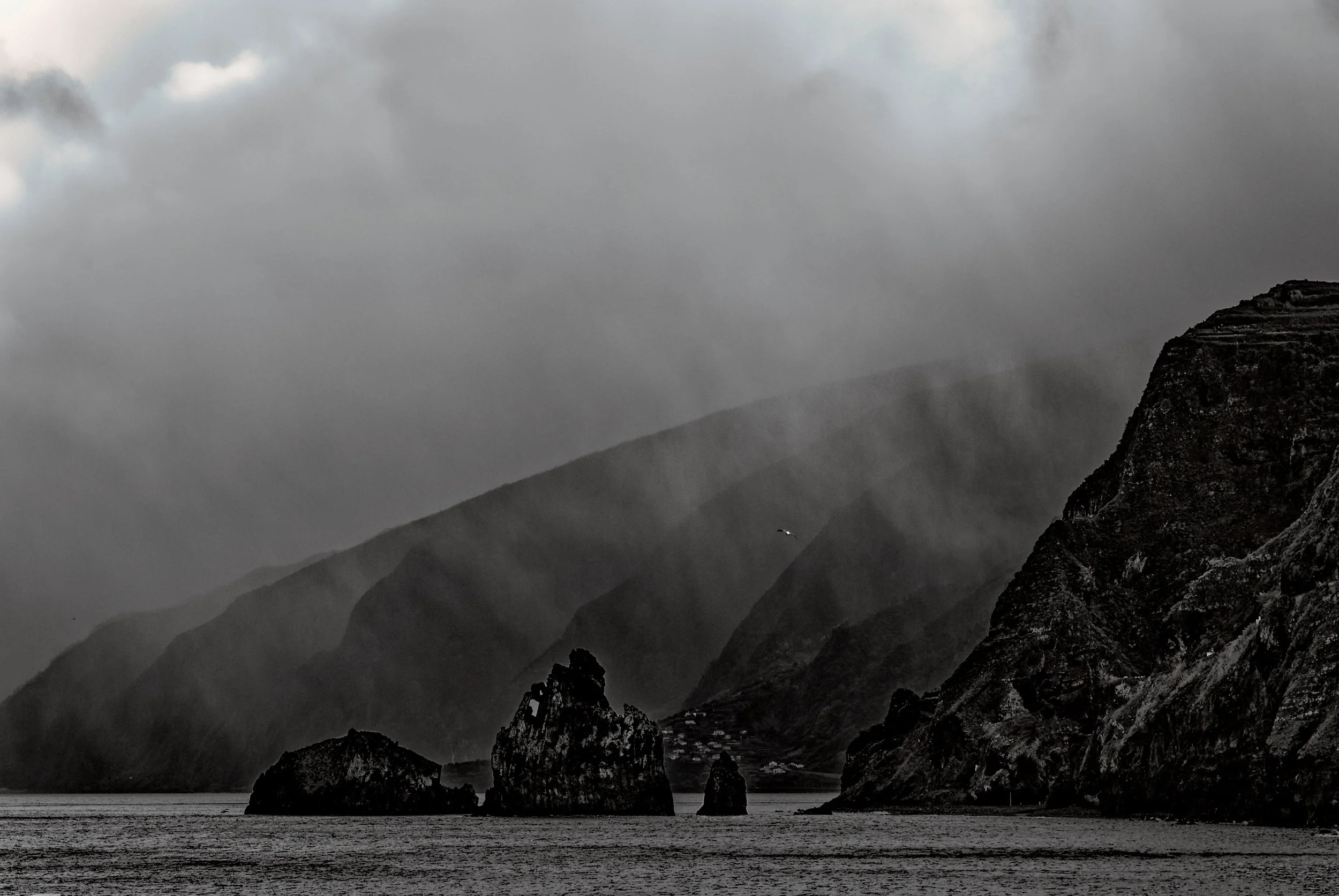 Dark, overcast mountain landscape with misty hills, rocky cliffs, a body of water in the foreground, and a bird flying over the water.