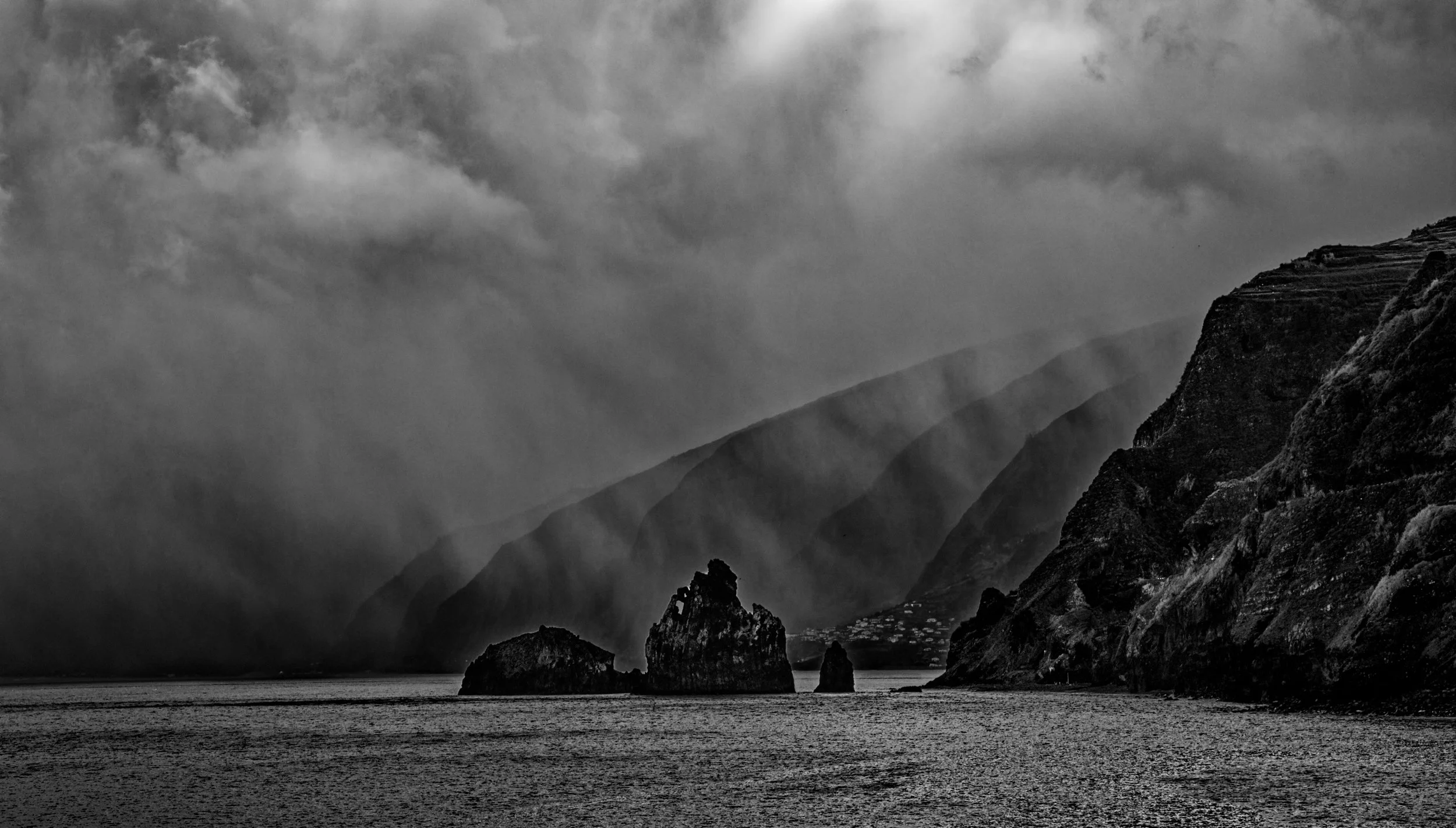 Black and white photograph of a mountainous landscape with steep cliffs, mist or clouds in the sky, and a body of water in the foreground.