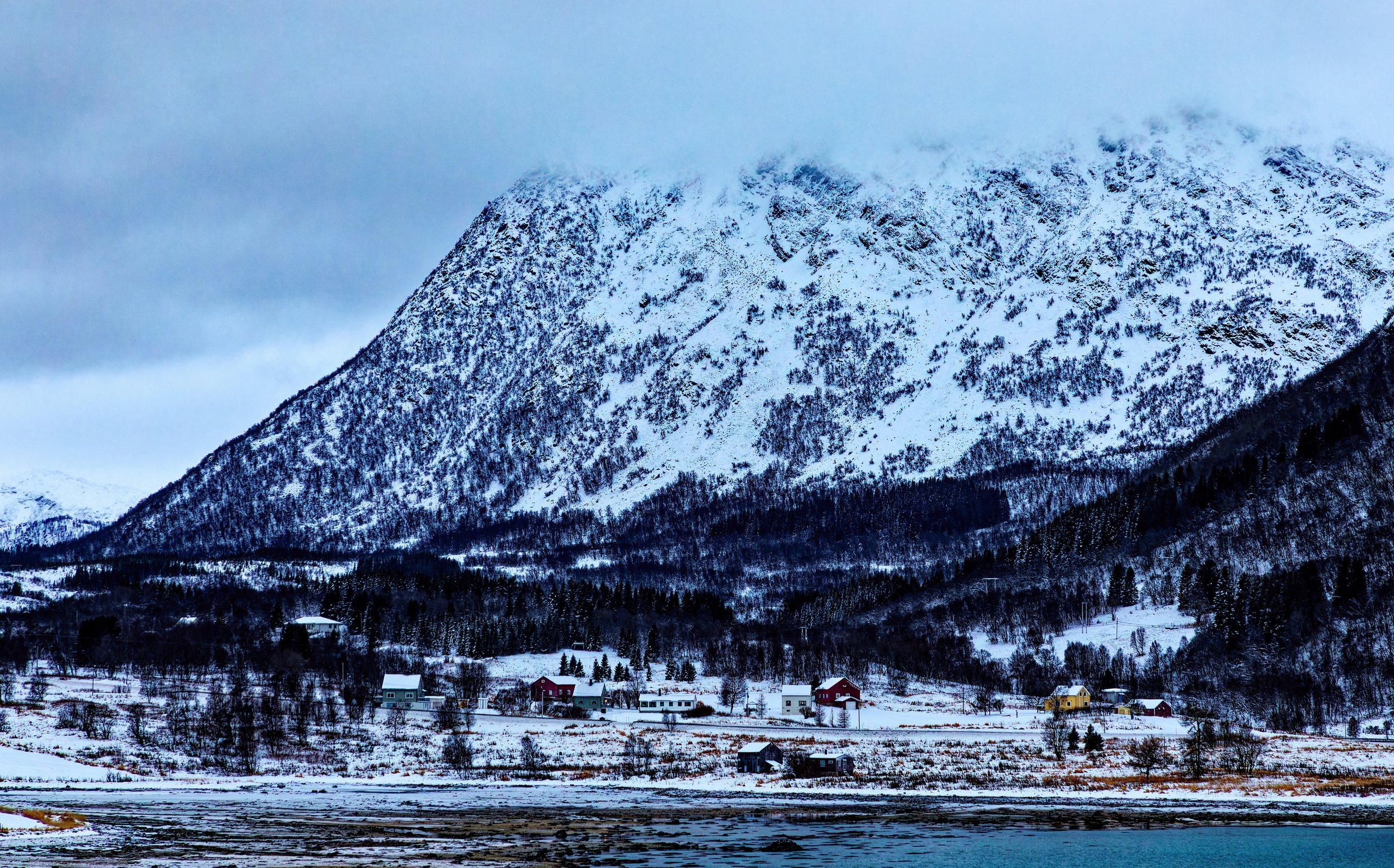 Snow-covered mountain with trees, low houses, and cloudy sky.