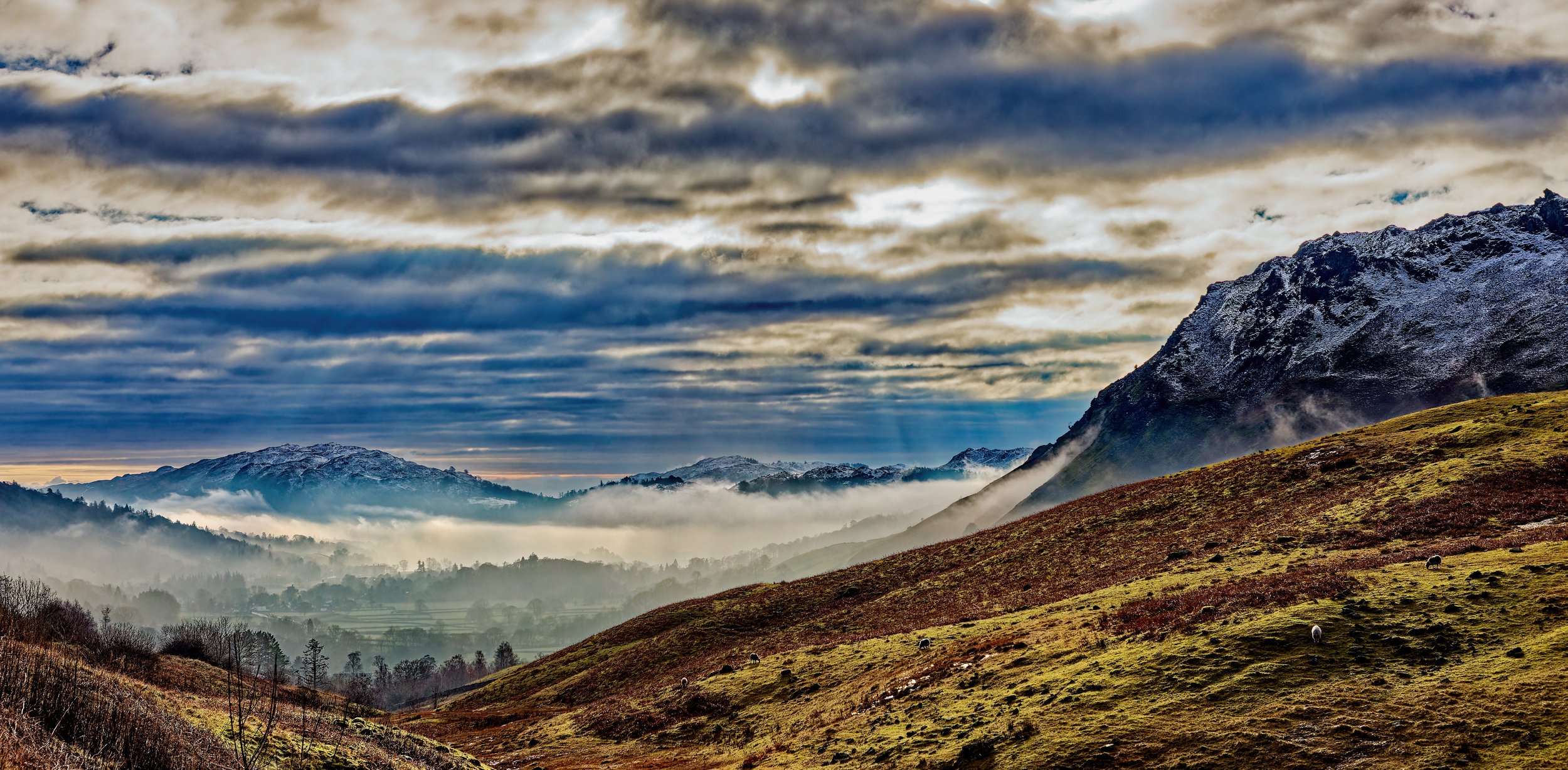 A scenic landscape of rolling hills and mountains, with some snow-capped peaks under a cloudy sky, and a valley filled with mist or low clouds, dotted with grazing sheep.