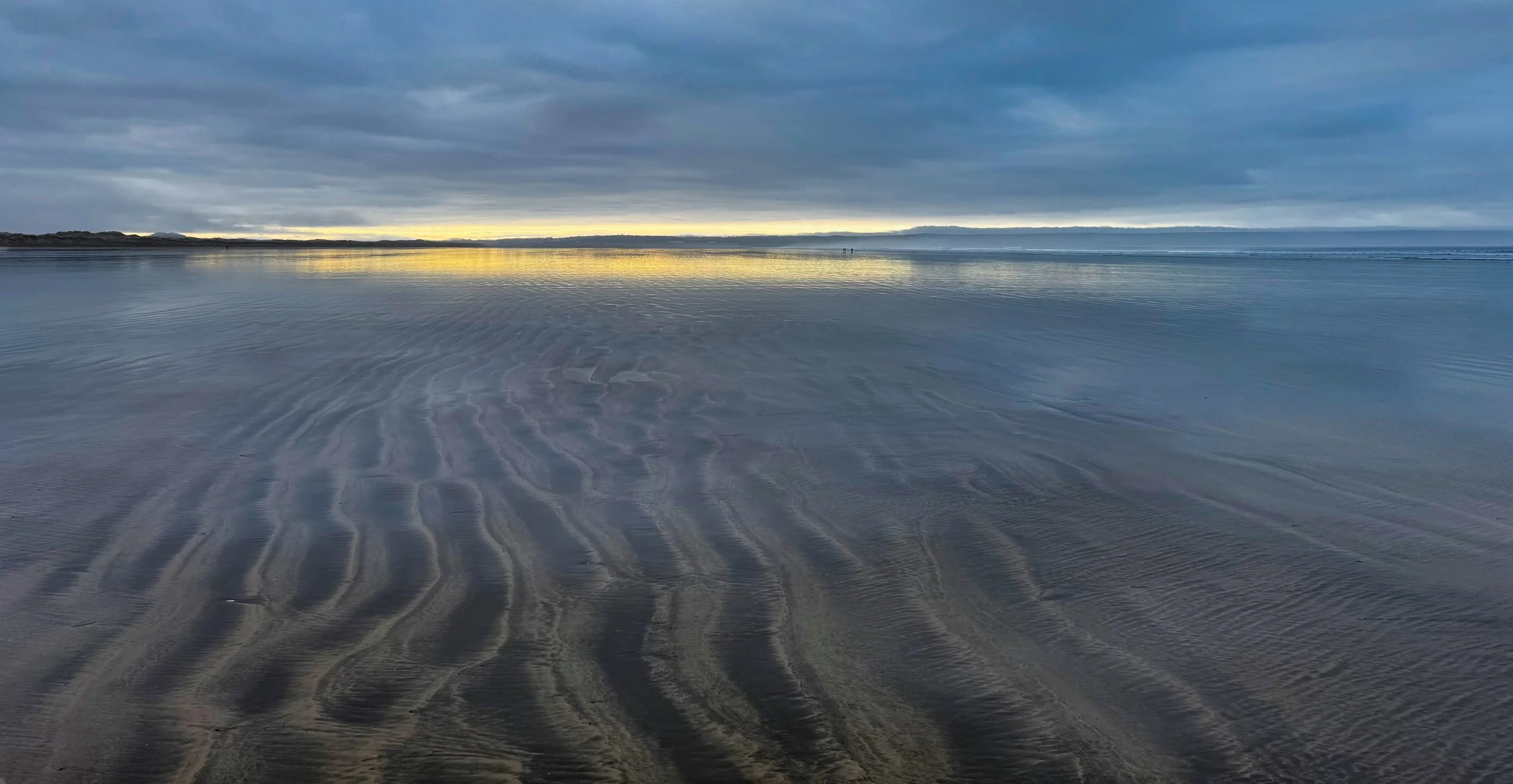 Overcast sky reflecting on a calm beach with rippled sand, distant shoreline, and smoke or fog on the horizon.
