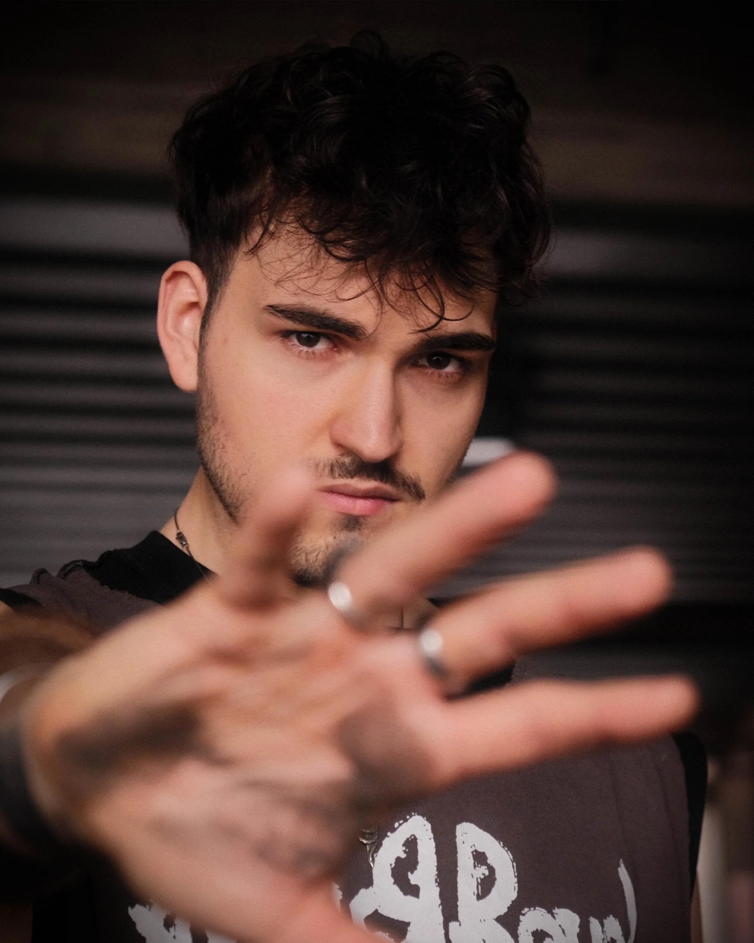 Ein junger Mann mit dunklen, lockigen Haaren und einem Bart blickt direkt in die Kamera. Im Hintergrund ist eine dunkle Wand mit horizontalen Linien sichtbar.