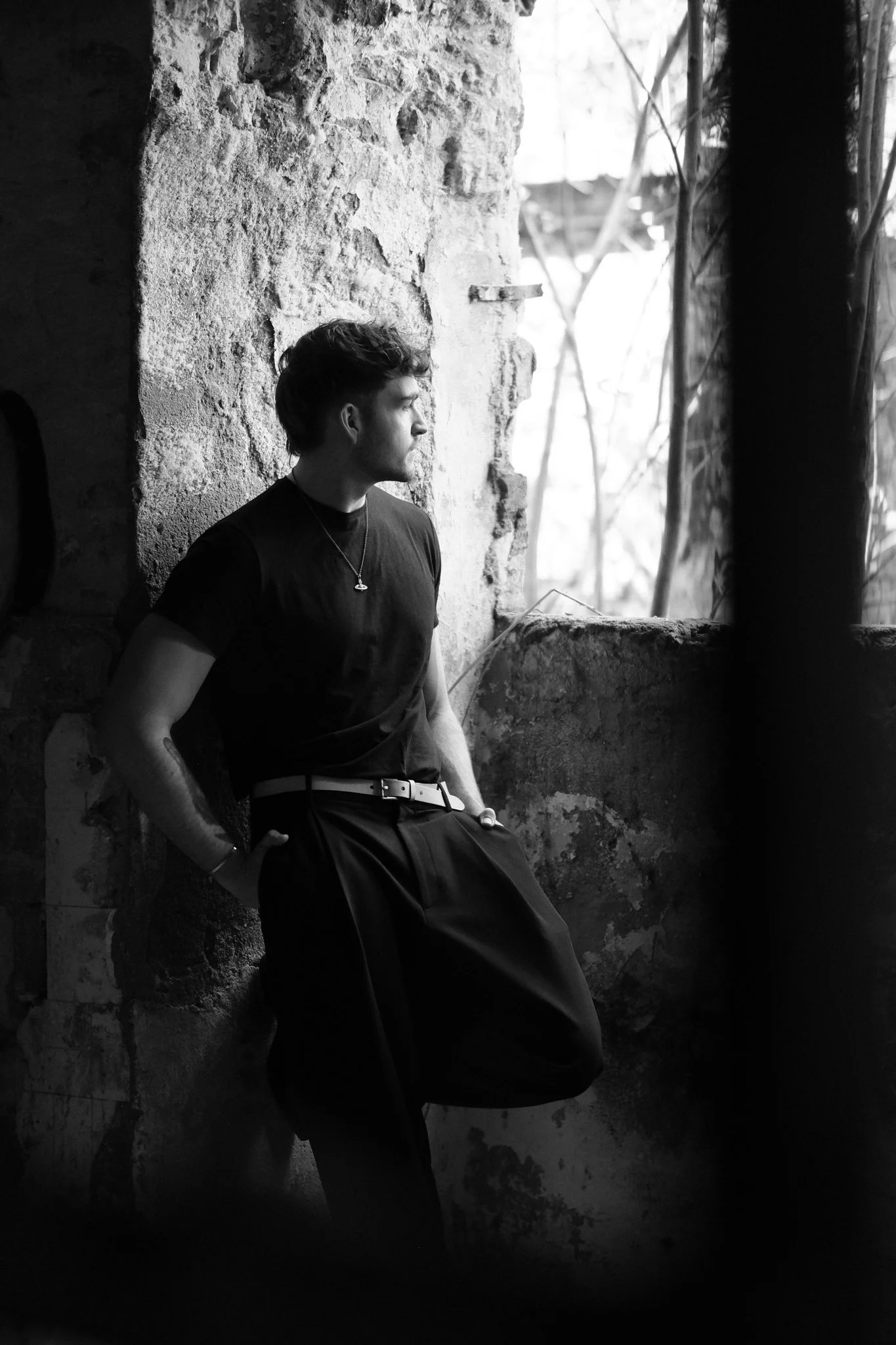 Schwarz-weiße Fotografie eines jungen Mannes, der entspannt an einer unordentlichen Ziegelmauer lehnt. Er trägt ein T-Shirt, eine Halskette und hat kurze, lockige Haare. Der Hintergrund zeigt eine offene Fensteröffnung mit Zweigen und Licht.