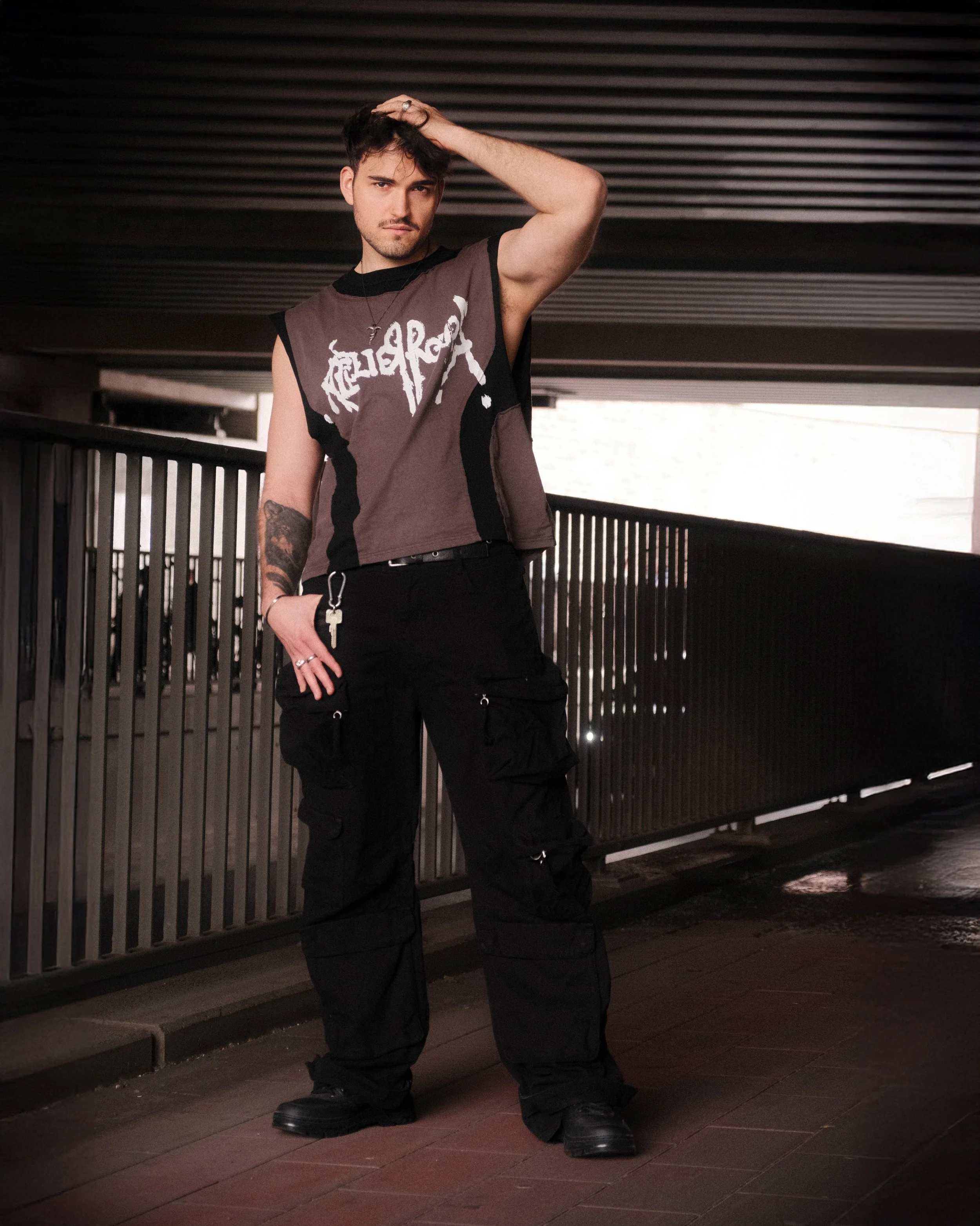 junger Mann mit dunklem Haar und Tattoos trägt ein zerstörtes Rockstar-T-Shirt, schwarze tactical-Hosen, Stehschuhe, im Schatten unter einer Brücke, schulterlang. Er steht am Bahnsteig mit einem Schlüsselbund in der linken Tasche, die rechte Hand auf