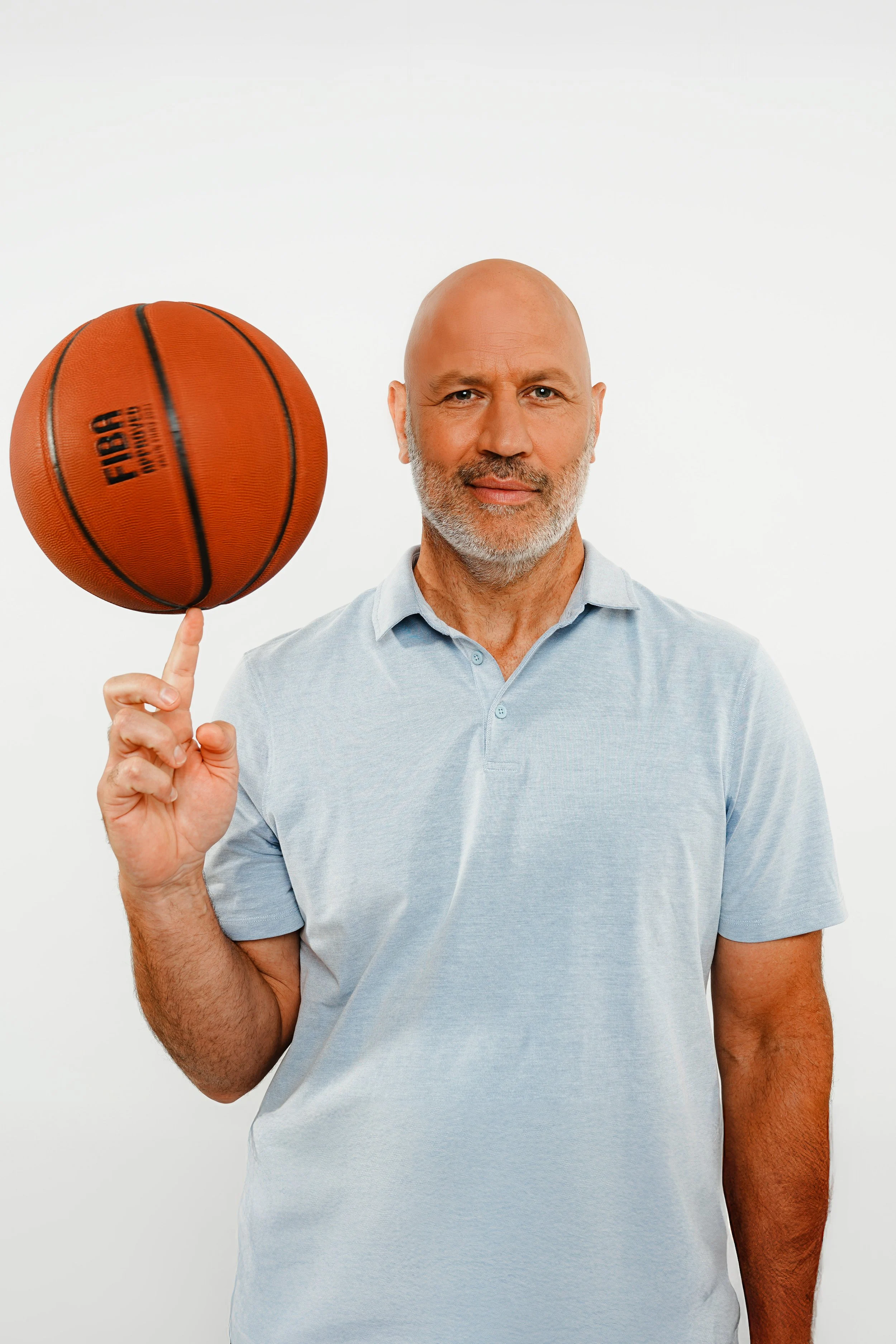 Ein älterer Mann mit Glatze und grauem Bart hält einen Basketball in der Hand vor einer weißen Wand.