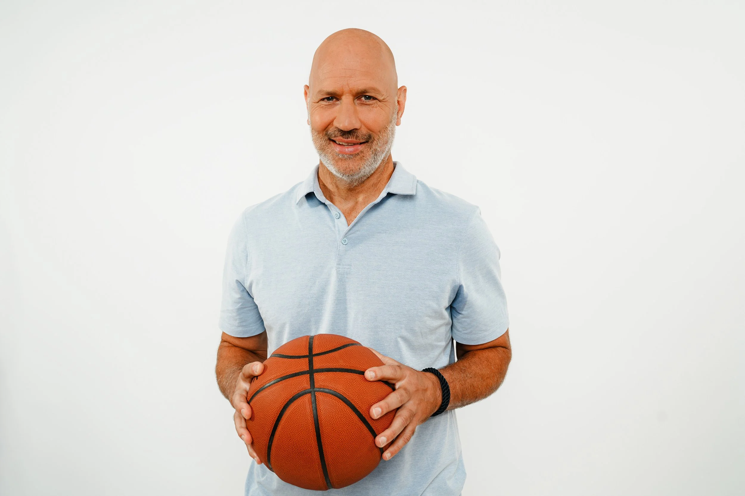 Ein älterer Mann mit Glatze und grauem Bart hält einen Basketball vor einem weißen Hintergrund.