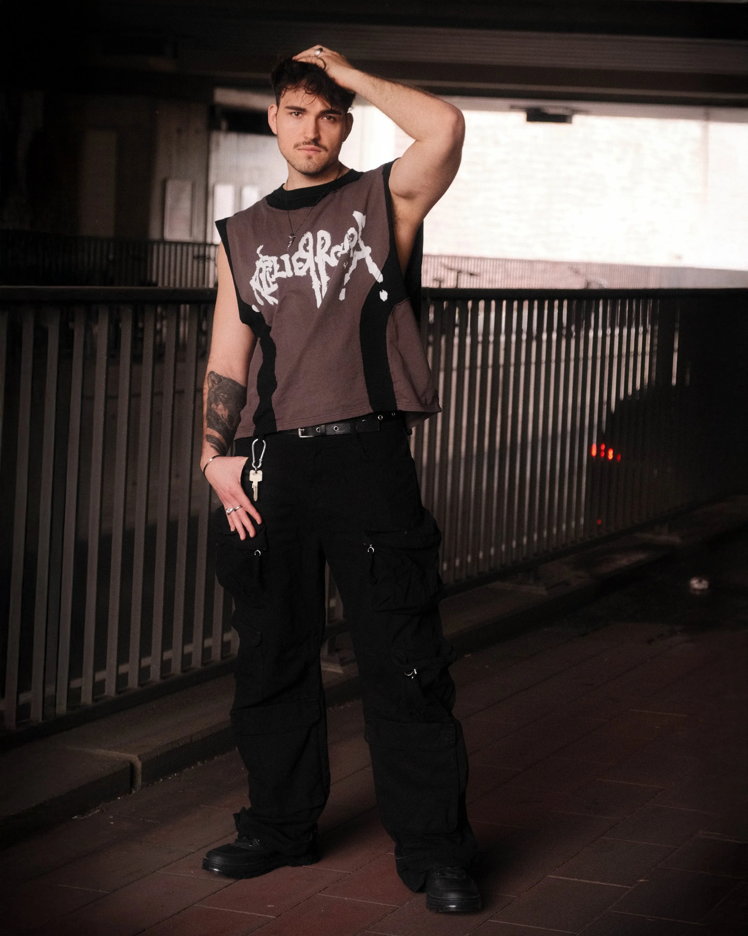 Junge Person mit schwarzen Haaren, tätowiertem Arm, trägt ein schwarzes und graues T-Shirt mit Print, schwarze Hose und Schuhe, steht in einem dunklen Raum mit Holzfußboden, Blick in die Kamera, Hand im Haar.