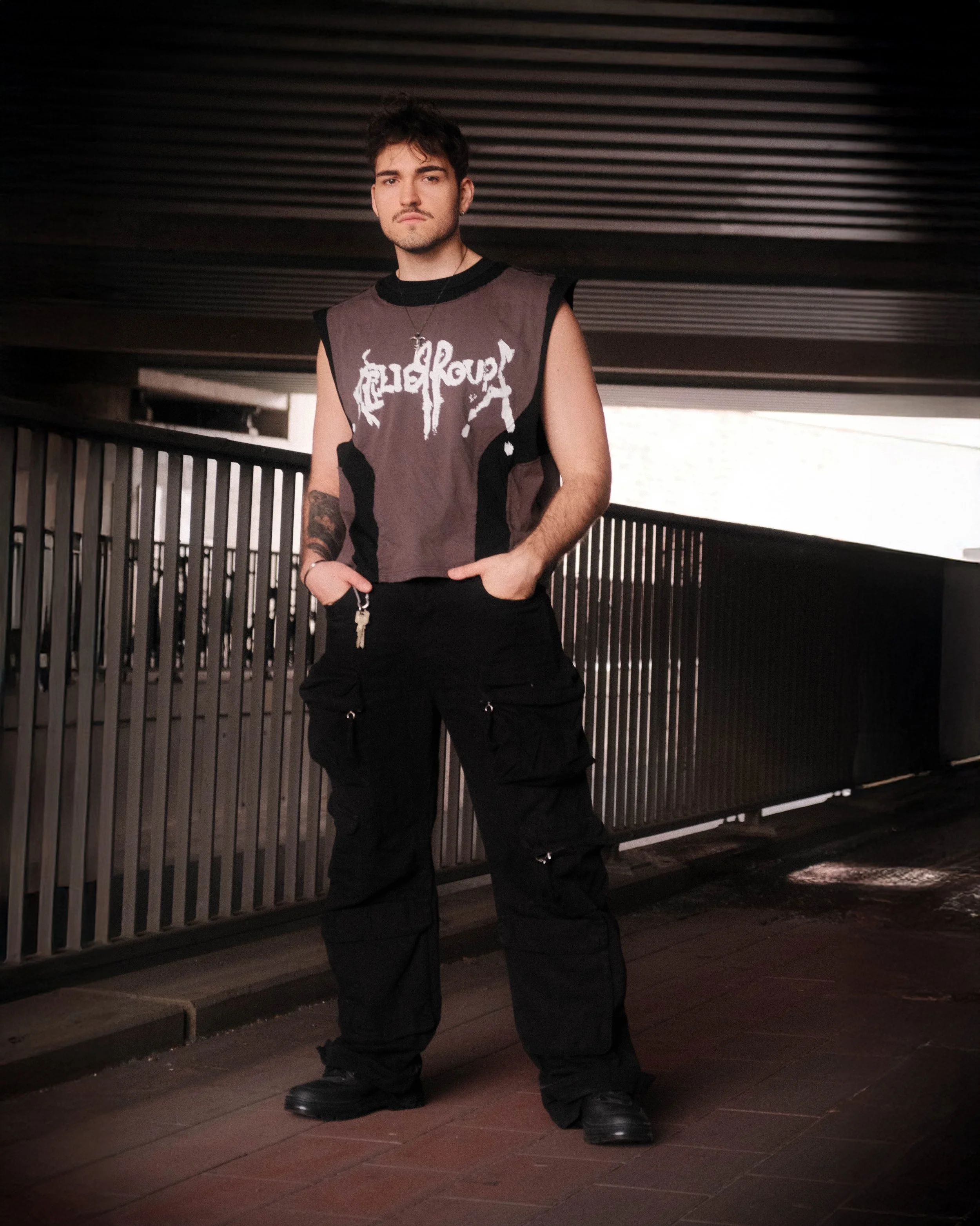 Junge Person steht vor einem Metallzaun auf einer dunklen Straße, trägt ein tönenfarbiges T-Shirt mit schwarzen Ärmeln und schwarzen Hosen, in einer urbanen Umgebung bei Nacht.