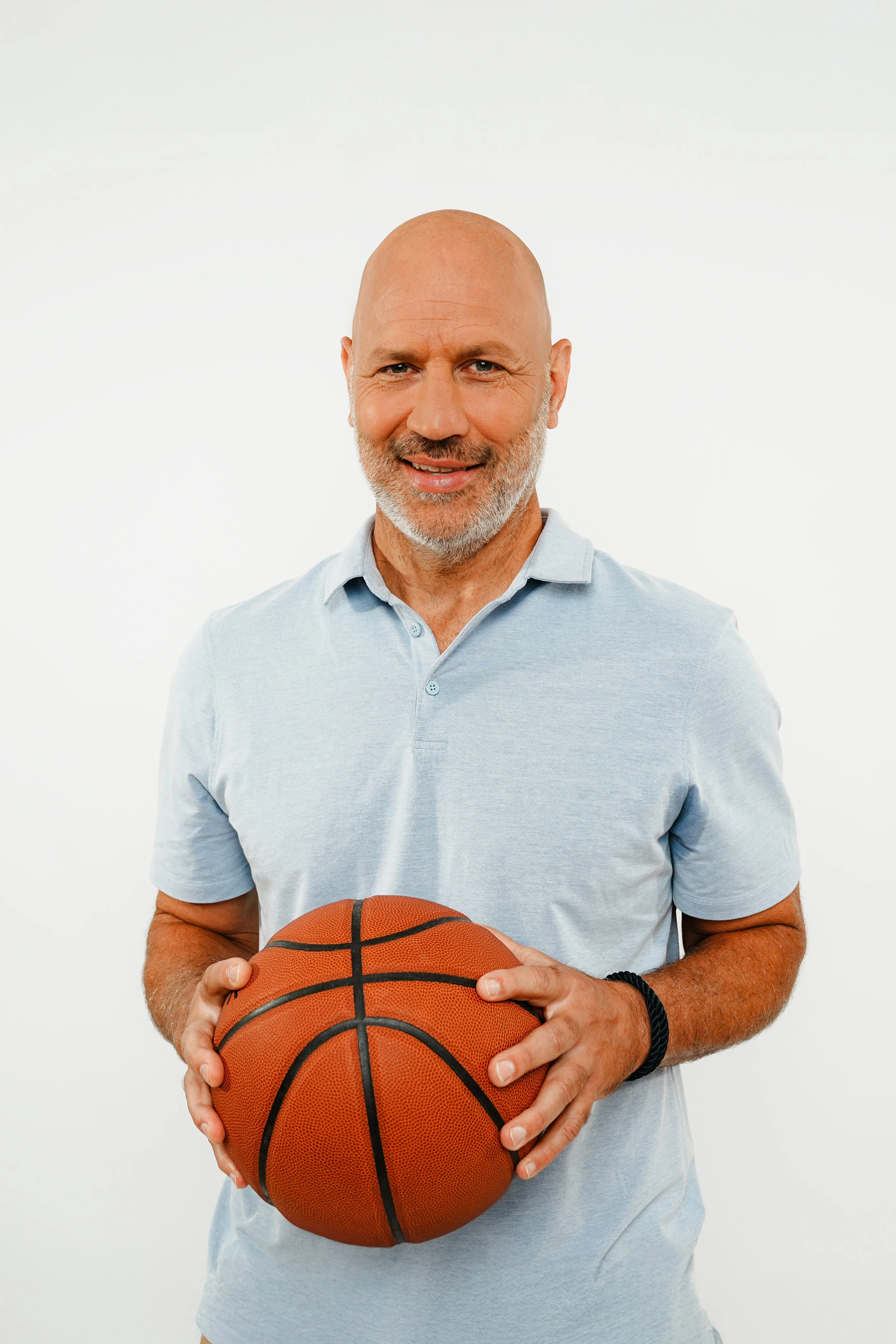 Älterer Mann mit sichtbarem weißen Bart, trägt hellblaues Polohem and hält Basketball in den Händen, steht vor weißen Hintergrund.