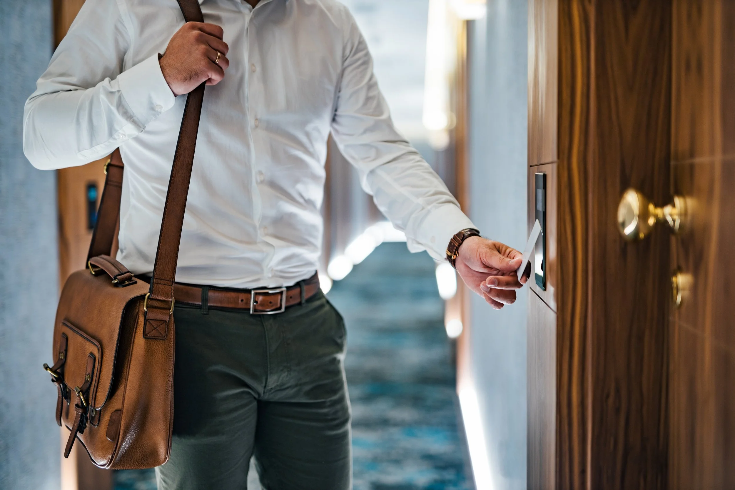 A man wearing a white shirt and dark pants is pressing a card against an electronic access panel, while carrying a brown leather shoulder bag.