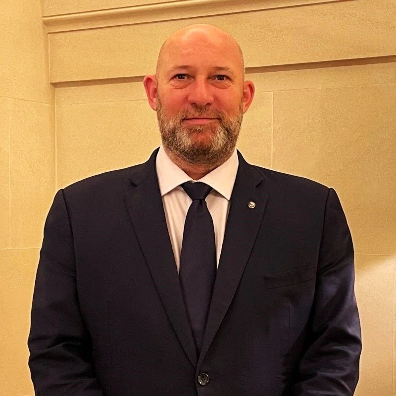 A man in a black suit with a white dress shirt and black tie standing against a beige wall.