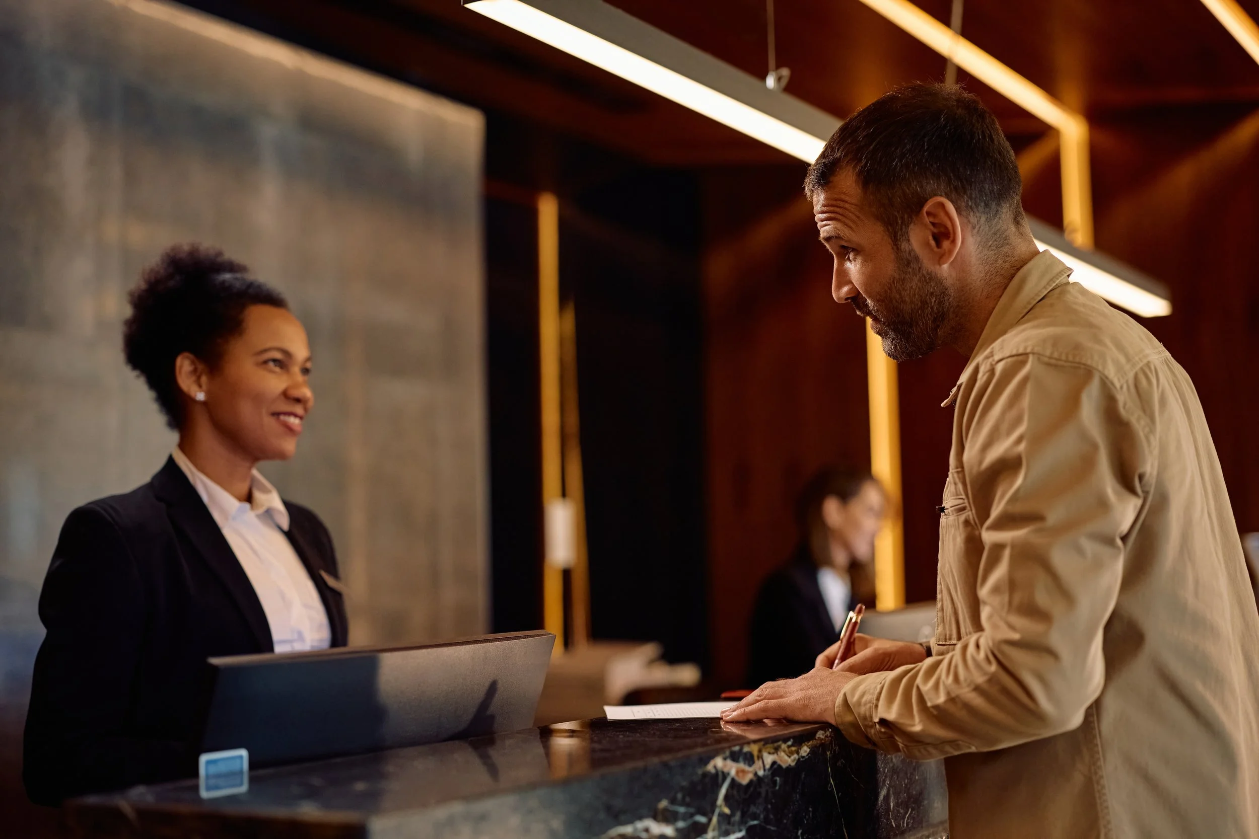 A hotel front desk clerk, an African American woman, smiling and assisting a man with a check-in, in a modern hotel lobby.