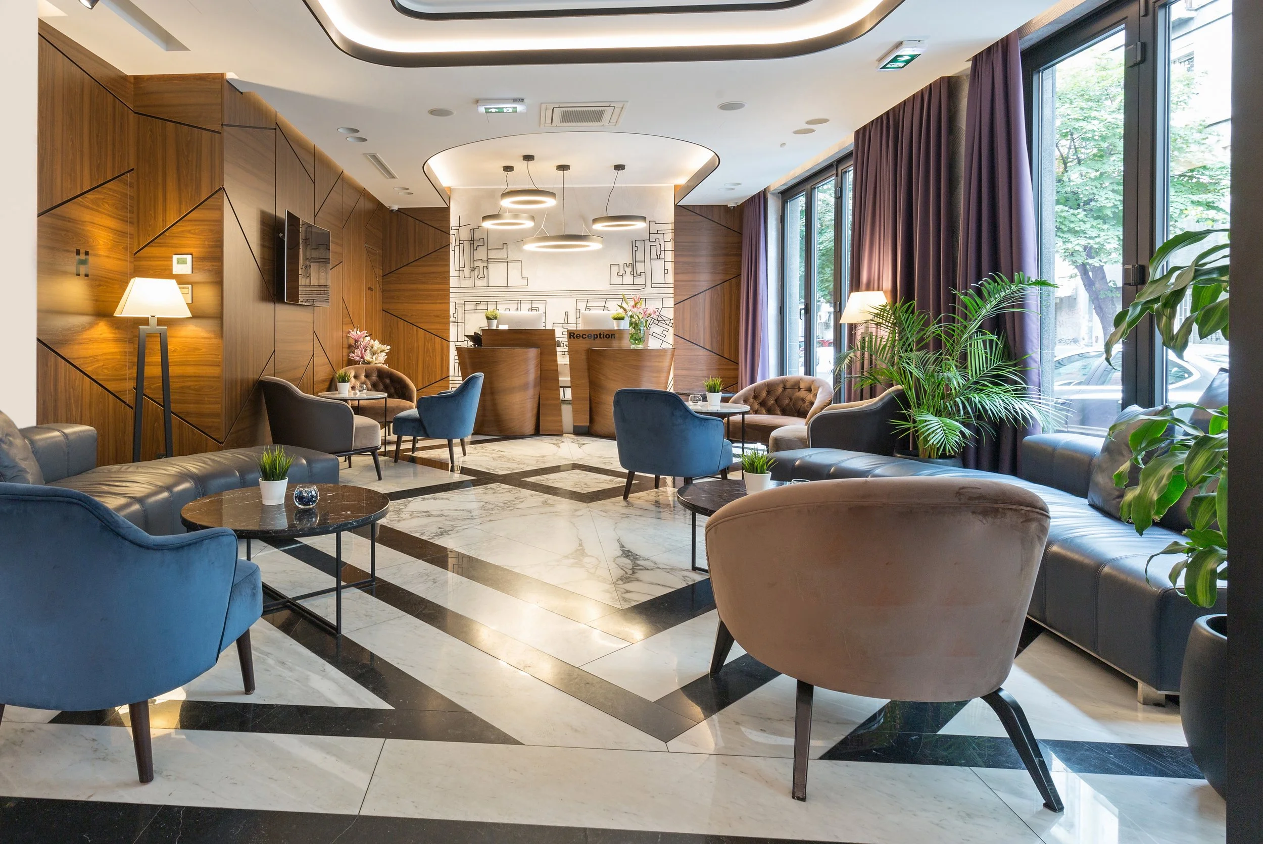 A hotel lobby with modern furniture, including blue and beige armchairs, small tables with potted plants, and a reception desk with flowers, illuminated by contemporary ceiling lights and large windows with purple curtains.