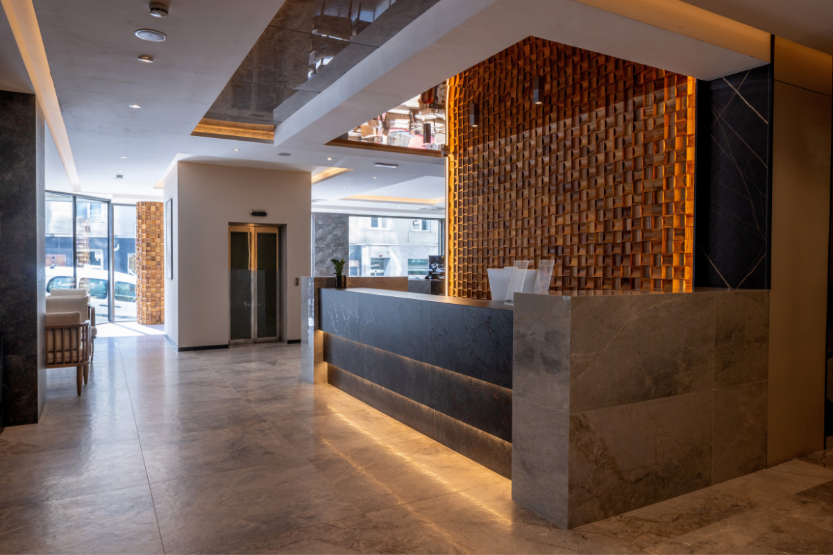 Modern hotel lobby with a reception desk, a decorative wooden wall, and seating area near large glass windows.